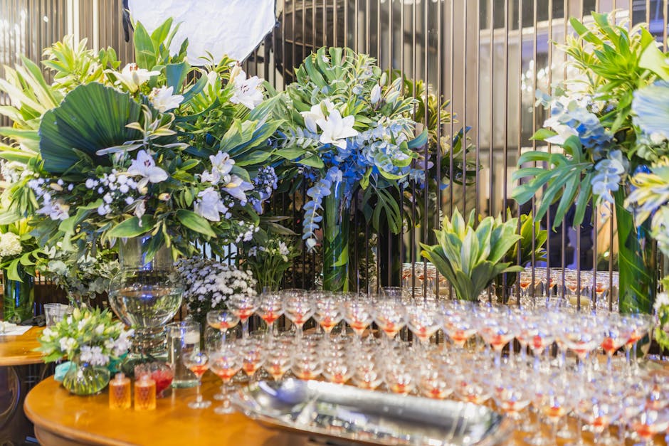 A floral arrangement featuring large tropical leaves, white lilies, and delicate white and purple flowers, displayed in clear glass vases on a wooden table. In front of the flowers, numerous small gla