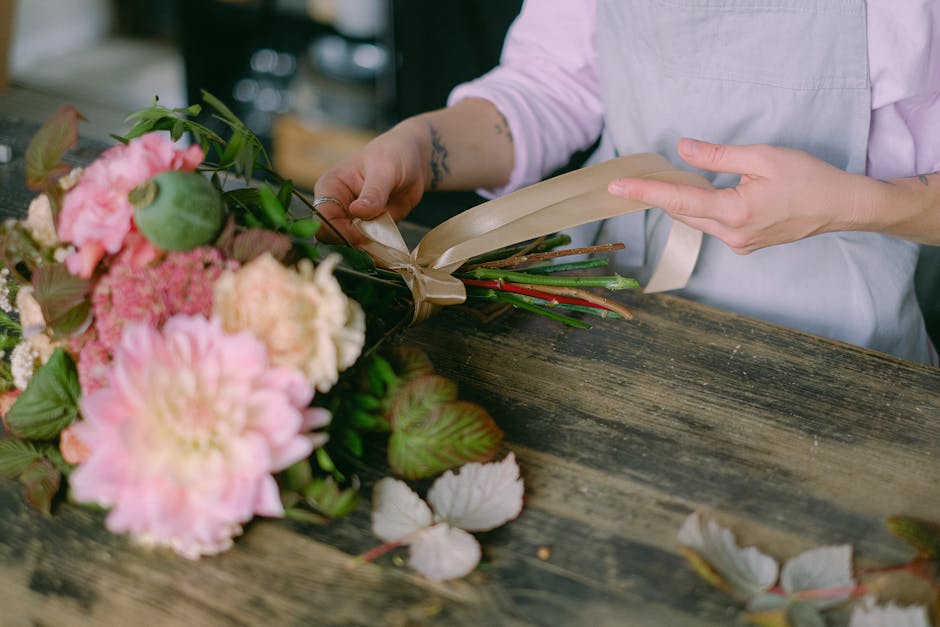A floral arrangement in progress on a rustic wooden table, featuring a bouquet of fresh pink peonies, hydrangeas, and greenery, with varying shades of pink and green. The florist, wearing a light pink