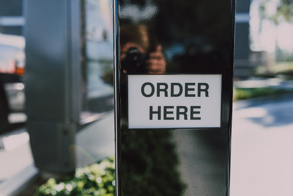A black outdoor kiosk with a white sign displaying 'ORDER HERE' in bold black letters, positioned on a sidewalk. In the background, blurry glimpses of parked cars and greenery are visible, with natura