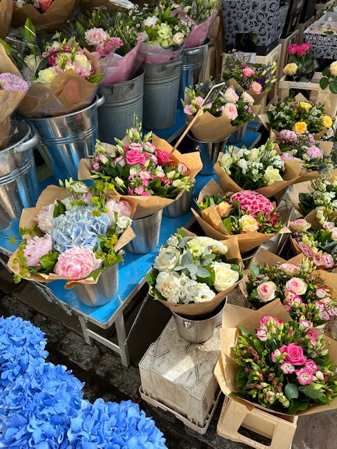 A vibrant display of fresh floral bouquets arranged in metal buckets and wrapped in brown kraft paper, featuring a variety of flowers including pink roses, white freesias, light pink blooms, and blue 