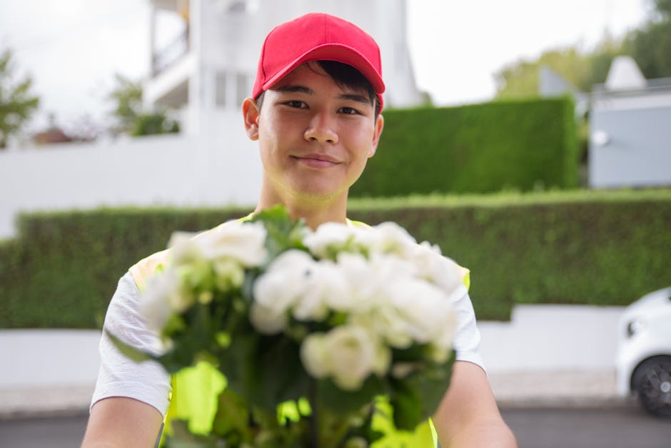 A delivery person wearing a red cap and a high-visibility vest is smiling while handing over a bouquet of fresh white flowers, including roses and other blooms, wrapped in clear cellophane. The bouque