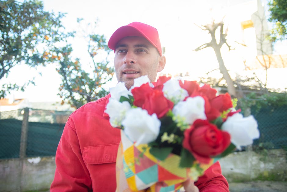 A man wearing a red cap and matching red jacket holds a vibrant bouquet of fresh flowers in front of him, consisting of red and white blooms, including roses and lisianthus, wrapped in colorful patter