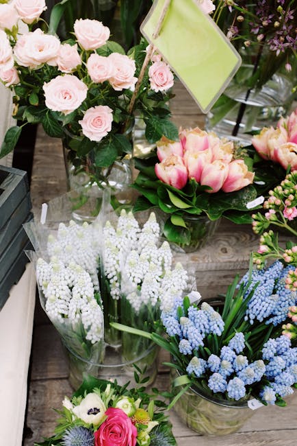A variety of fresh floral arrangements displayed on a wooden table, including a bouquet of light pink roses with dark green foliage in a clear glass vase, a cluster of pink tulips with green leaves, a
