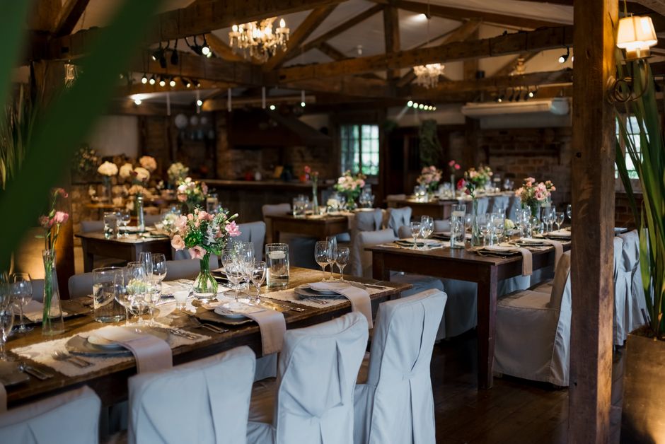 Elegant wedding table setting in a rustic venue with exposed wooden beams and soft warm lighting. The tables are decorated with fresh flower arrangements in glass vases, featuring pink, white, and cre