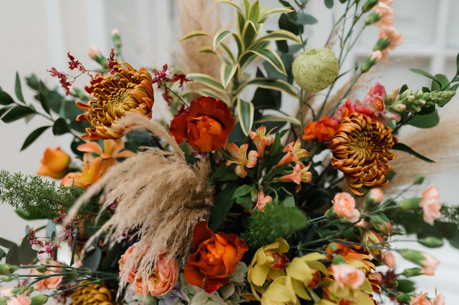Close-up of a floral arrangement featuring a variety of fresh flowers including orange and yellow roses, large brownish-orange chrysanthemum blooms, delicate pink and peach sprays, and green foliage w